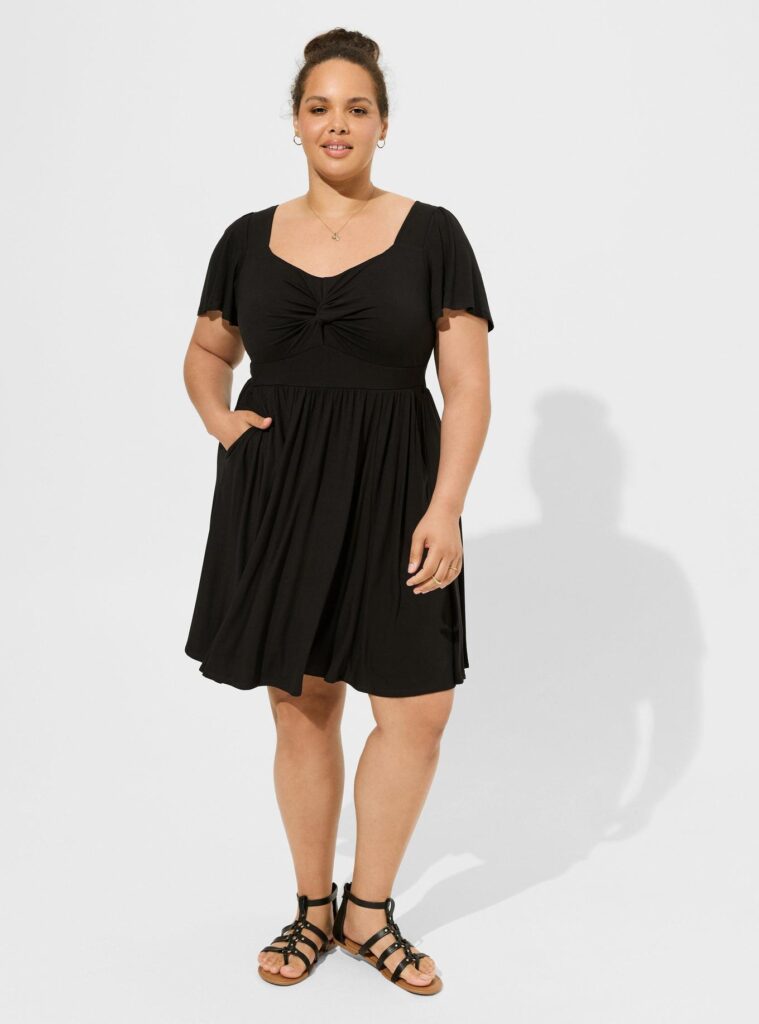 A woman wearing a black dress, necklace, sandals and earrings. The dress is black with flutter sleeves and a tie in the front.