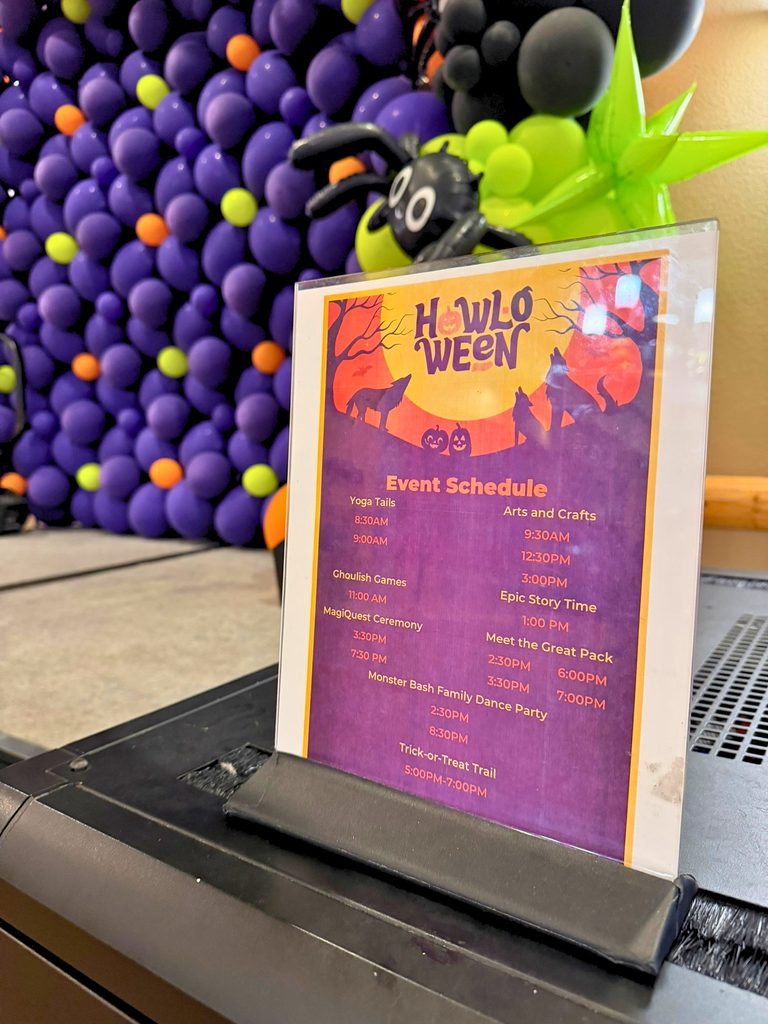 Great Wolf Lodge Howl-O-Ween 2026 Event Schedule sign in front of a stage with a purple Halloween balloon wall. Must Do Halloween Activities for Kids in Southern California: Great Wolf Lodge Garden Grove Howl-O-Ween